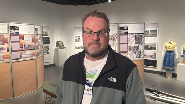 Portrait en plan moyen d'un homme portant des lunettes et une veste zippée, debout au centre d'une galerie d'exposition entouré de grands panneaux explicatifs avec photos d'archives et d'un mannequin costumé.