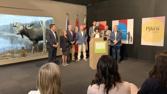 Ginette Petitpas Taylor s'adresse à une petite foule dans un musée. Un orignal empaillé est à sa droite.