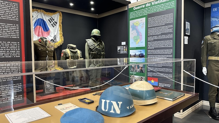 Des casques de soldats sont déposés sur une table d’exposition protégée par des vitres devant des panneaux explicatifs et des uniformes de soldat.