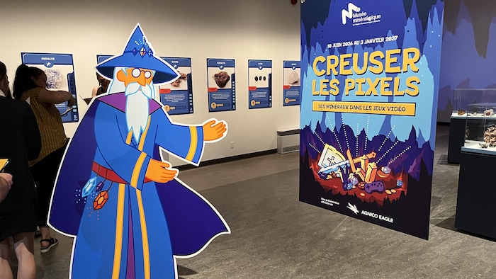 Une affiche en carton grandeur nature d'un personnage de sorcier qui pointe vers l'affiche de l'exposition Creuser les pixels.