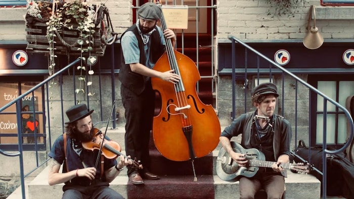 Three men play music on outdoor steps. On the left, a bearded man wearing a hat plays the violin. In the center, a bearded man with a cap plays double bass. On the right, a man wearing a hat plays an acoustic guitar. They are in front of a brick building and flower pots. street musicians forced sing french