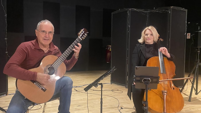 Un duo de musiciens est assis sur une scène en bois. À gauche, un homme d'âge mûr joue de la guitare classique. À droite, une femme aux cheveux blonds clairs joue du violoncelle, avec une tablette musicale numérique montée devant elle. L'arrière-plan est sombre, avec des séparateurs noirs.