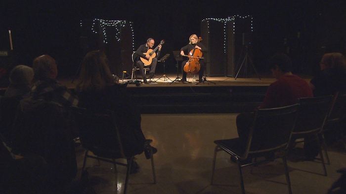 Un duo de musiciens, composé d'un guitariste et d'une violoncelliste, joue sur une petite scène éclairée par le haut. Quelques guirlandes lumineuses décorent l'arrière-plan sombre. Le public est assis dans le noir au premier plan, regardant la performance.