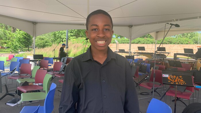 Un adolescent membre d'un orchestre debout sous un chapiteau. Il regarde la caméra et sourit.