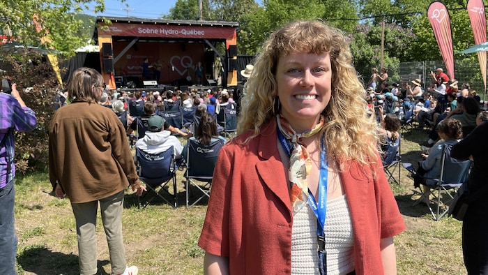 Une femme souriante aux cheveux bouclés pose devant une foule et une scène extérieure lors d'un festival en plein air.
