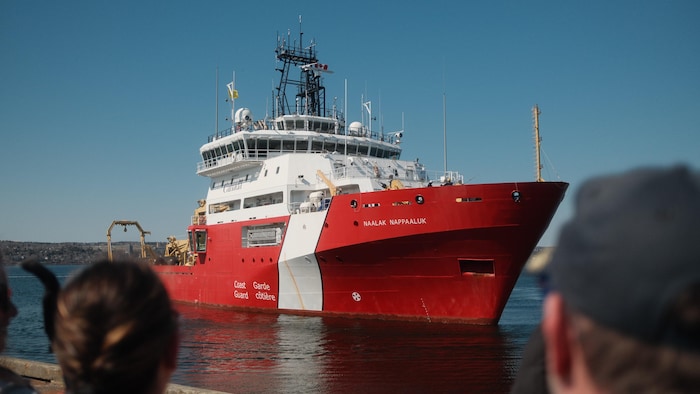 Quelques personnes regardent un gros navire rouge et blanc s'approcher à quelques mètres du quai sous un ciel bleu sans nuage.