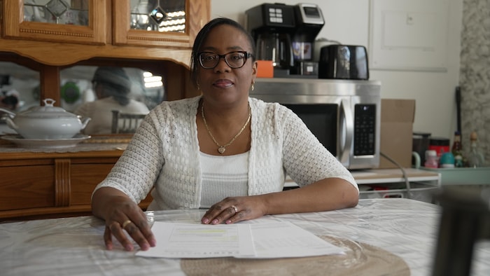 Une femme à sa table de cuisine regarde dans la caméra. Des papiers sont sur la table devant elle.