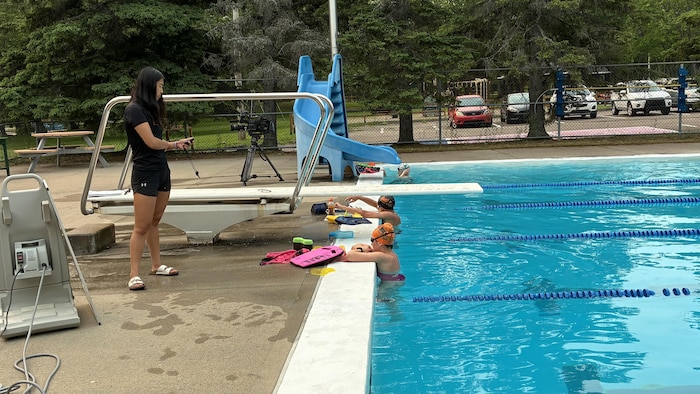 Deux nageurs s'entraînent dans la piscine sous la supervision de leur entraîneuse, postée à l'extérieur.
