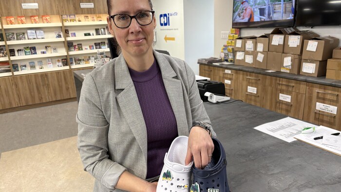 Nancy Arpin pose pour la caméra en souriant légèrement, tout en tenant une paire de sandales sur lesquelles sont accrochées des breloques.