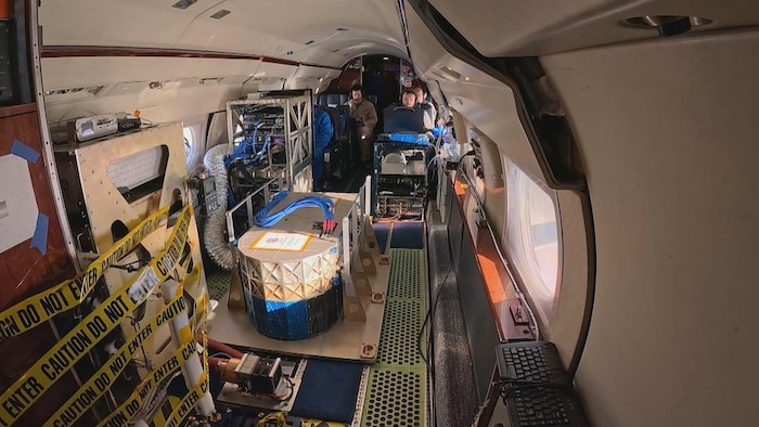 L'intérieur d'un avion rempli d'instruments et de fils. Quatre personnes au fond sont assises sur des bancs.