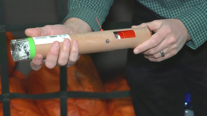 Un cylindre brun tenu par deux mains d'homme. Une ouverture rectangulaire laisse entrevoir un objet rouge à l'intérieur.