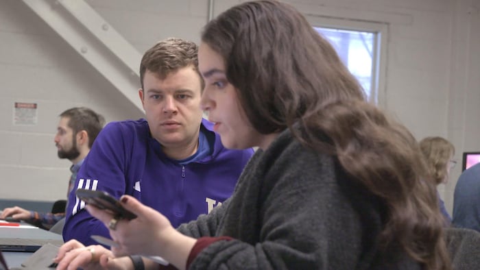 Une femme de profil examinant un téléphone et tapant sur un clavier. Un homme la regarde d'un air concentré. D'autres personnes sont assises derrière.