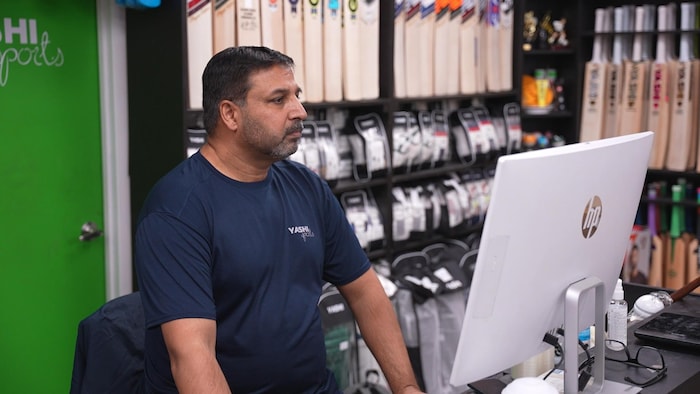 Nasir Mahmood regarde un écran d'ordinateur dans un magasin où on aperçoit des gants et des bâtons de cricket.