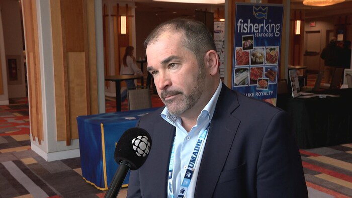 Profil gauche de Nat Richard, affichant un air sérieux, un micro devant lui, dans une salle de conférence.