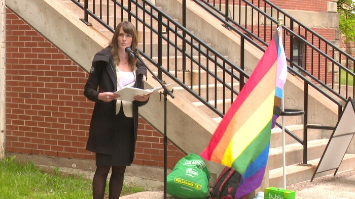 Natalie Jameson, debout avec une feuille à la main, parle au micro à côté des escaliers d'un édifice et près d'un drapeau arc-en-ciel.