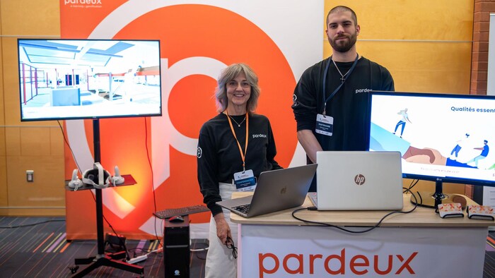 Une femme et un homme debout dans un stand avec deux ordinateurs portables et deux écrans de présentation