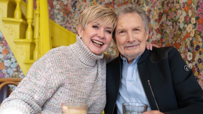 Une femme blonde et un homme aux cheveux gris sourient. 