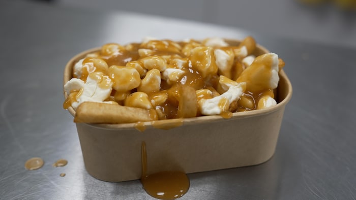 Une poutine généreuse, composée de frites, de gros grains de fromage en crottes et de sauce brune coulante, est servie dans un contenant de carton brun. Le plat repose sur une surface métallique brossée avec quelques gouttes de sauce.