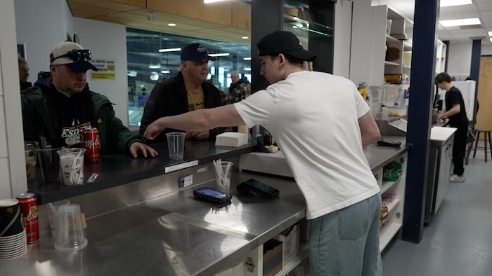 Un jeune employé sert deux clients au comptoir d'un casse-croûte situé dans un aréna. Des canettes de bière, des verres en plastique et un terminal de paiement sont visibles sur la surface de service en inox.