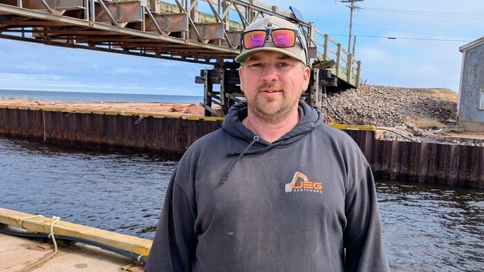 Un homme debout sur un quai, près d'un pont en bois. Il a des lunettes de soleil roses sur sa casquette.