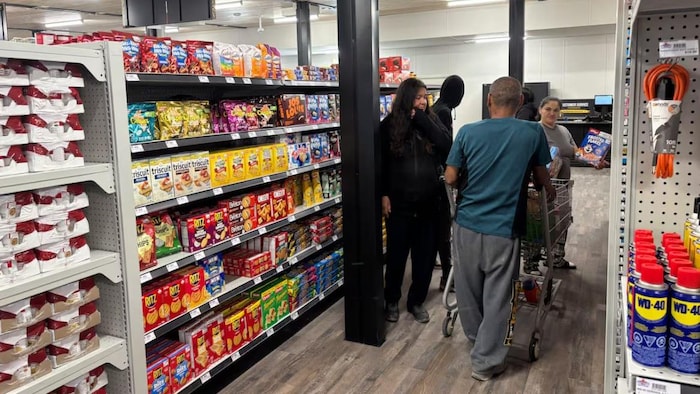 Des clients font leurs courses dans les allées d'une épicerie, où les panneaux indicateurs suspendus affichent les catégories de produits en anglais et en langue autochtone.