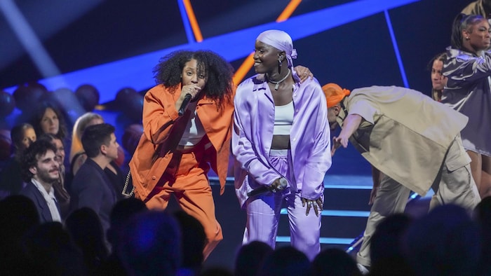 Deux femmes, l'une en orange et l'autre en mauve, chantent sur scène. 