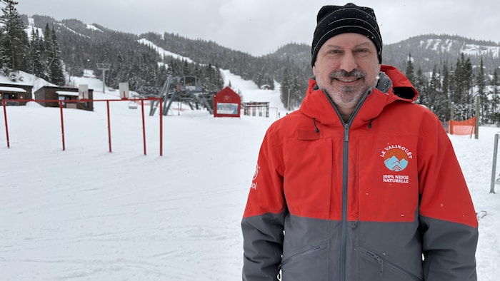 Un homme en manteau sourit à la caméra devant un décor de montagne de ski.
