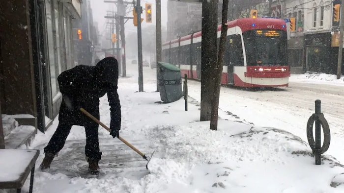شخص يقوم بإزالة الثلوج عن أحد الأرصفة في تورونتو فيما يمر قطار ترامواي في الشارع.