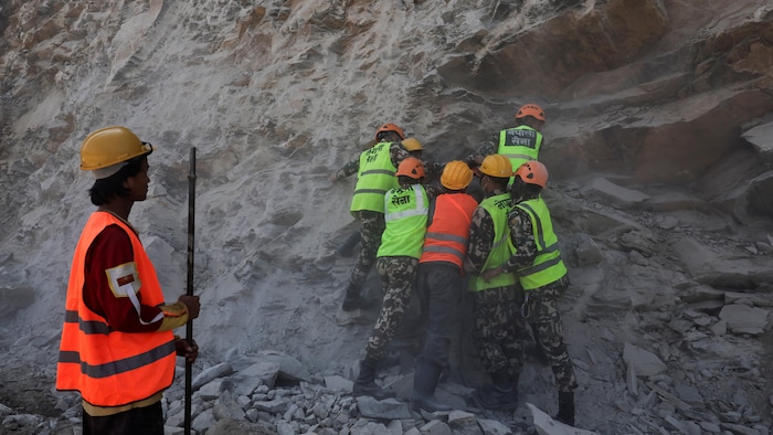 Des hommes vêtus d'un treillis militaire et de gilets orange et jaune devant une falaise.