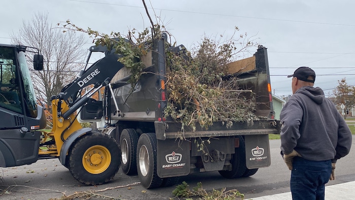 Un homme sur le trottoir regarde une pelle mécanique remplir un camion de branches d'arbres.