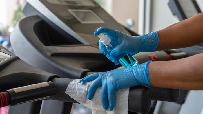 Une employée, les mains protégées par des gants bleus, applique du désinfectant sur les poignées d'une machine d'exercice dans une salle de sport.