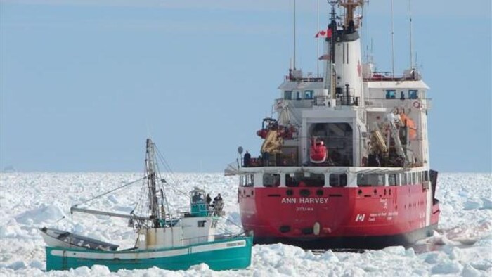 Un navire de la Garde côtière canadienne, l'Ann Harvey, est sur l'eau à travers les glaces et s'approche d'un petit bateau de pêche turquoise.