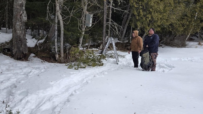 Deux hommes debout dans la neige avec une petite échelle et un sac à dos, devant un arbre sur lequel est accroché un nichoir.