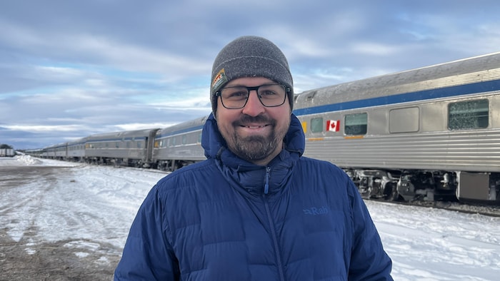 Un homme avec des lunettes et une tuque devant un train déraillé.