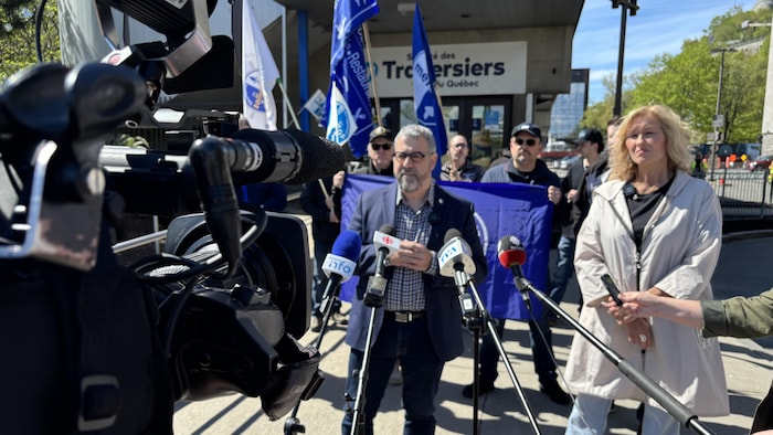 Nicolas Lapierre et Magali Picard lors d’une mêlée de presse à l’extérieur. Derrière eux, des syndiqués brandissent des drapeaux aux couleurs des Métallos.