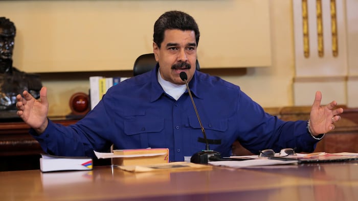Un homme qui a des cheveux noirs courts qui commencent à grisonner et qui a une moustache noire parle dans un micro. Il est vêtu d'une blouse bleue et porte un t-shirt blanc en dessous.