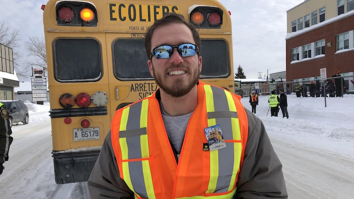 Un homme portant une veste fluo sourit à la caméra devant un autobus scolaire.