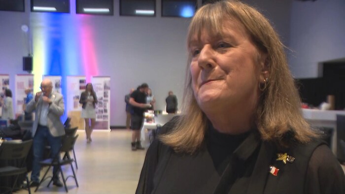 Une femme se positionne pour une entrevue à la caméra tandis qu’un homme s’empiffre à l’arrière.