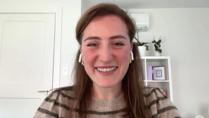 Une femme devant sa webcam sourit. Elle a des écouteurs sans-fil dans les oreilles et est dans une pièce aux murs blancs, devant une étagère décorée de plantes.