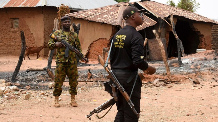 Des hommes armés patrouillent dans un village devant une maison brûlée.