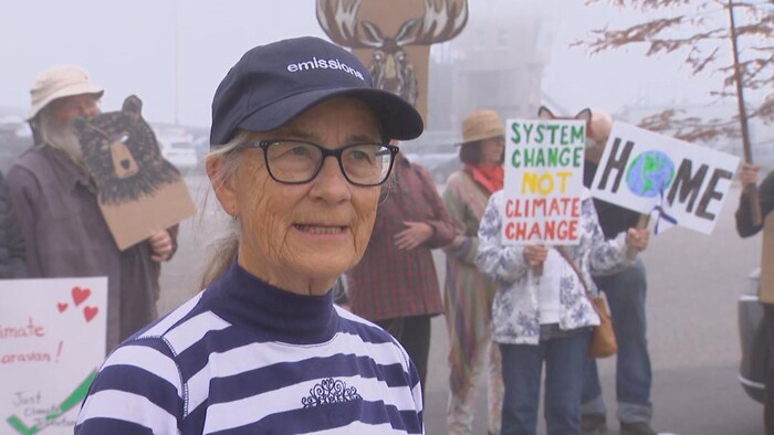 Une aînée porte une casquette et des lunettes. Elle est devant un regroupement de gens qui s'inquiètent des changements climatiques.