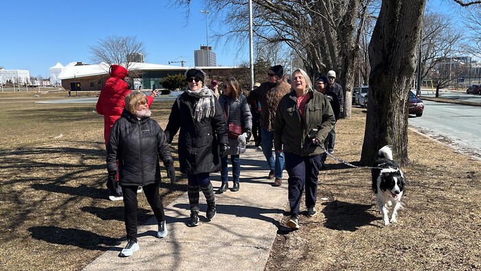 Un groupe mené par trois femmes, dont une tient un chien en laisse, avance sur un trottoir, le long d'un parc.