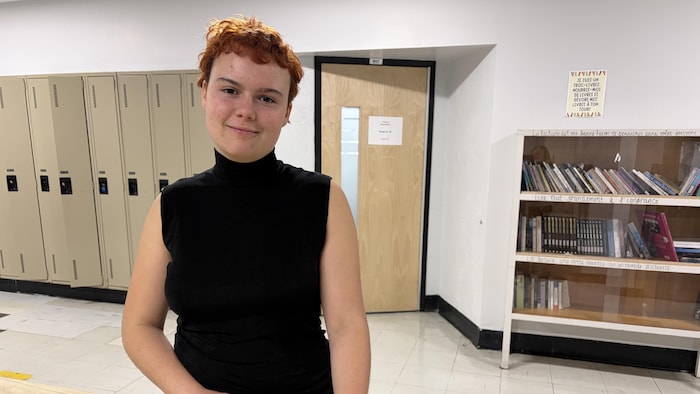 Une jeune femme se tient debout derrière une table, avec des casiers et une bibliothèque derrière elle.