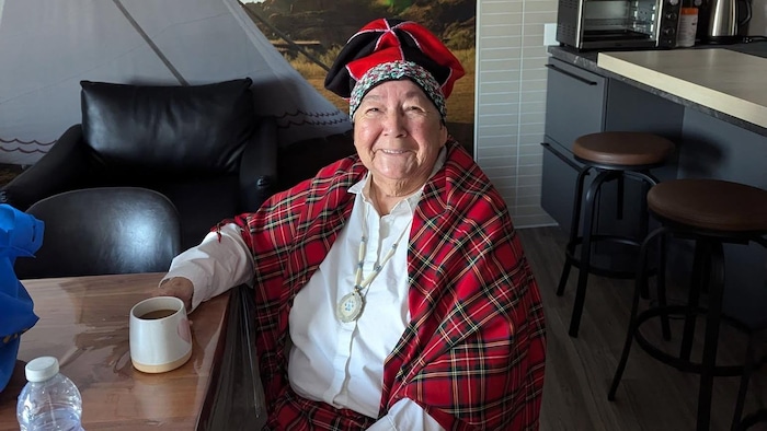 Noella Mckenzie est assise en tenue traditionnelle et sourit à la caméra, une tasse de café à la main