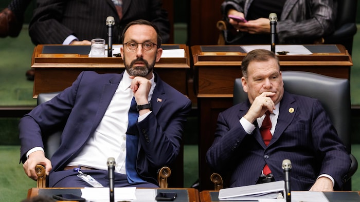 Le ministre Nolan Quinn assis à son siège en chambre à côté du ministre Todd McCarthy.