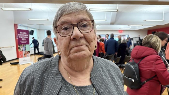 Une femme sourit à la caméra, des gens derrière elle dans une salle.