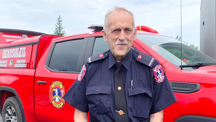Un pompier, en uniforme bleu foncé et cravate noire, est debout devant un véhicule rouge du service d'incendie de Beausoleil.