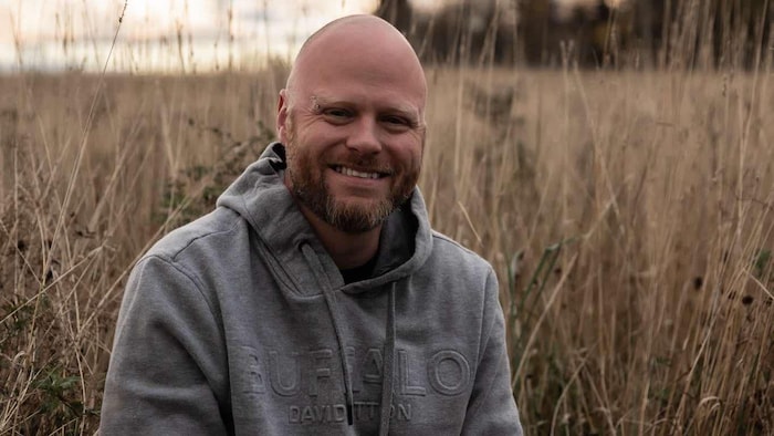 Un homme sourit à la caméra, dans un champ.