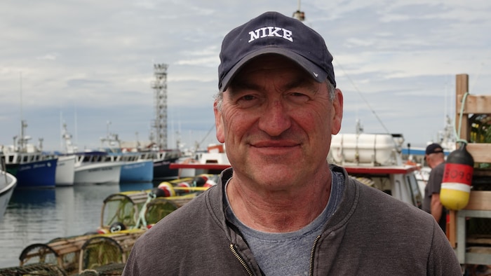 Un homme regarde l'objectif avec un sourire, il est devant un port de petits bateaux.