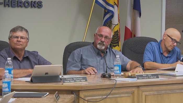 Le maire est assis à la table du conseil municipal entre deux conseillers.